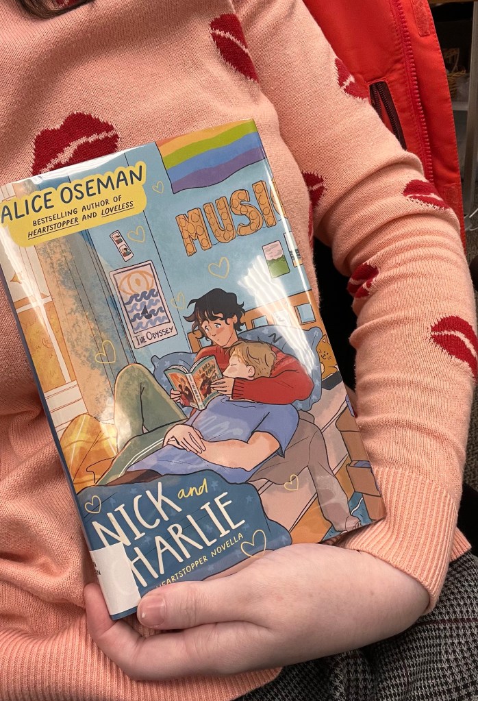 A photo of the novella, "Nick and Charlie" by Alice Oseman being held by a woman in a pink sweater with red lipstick prints all over
